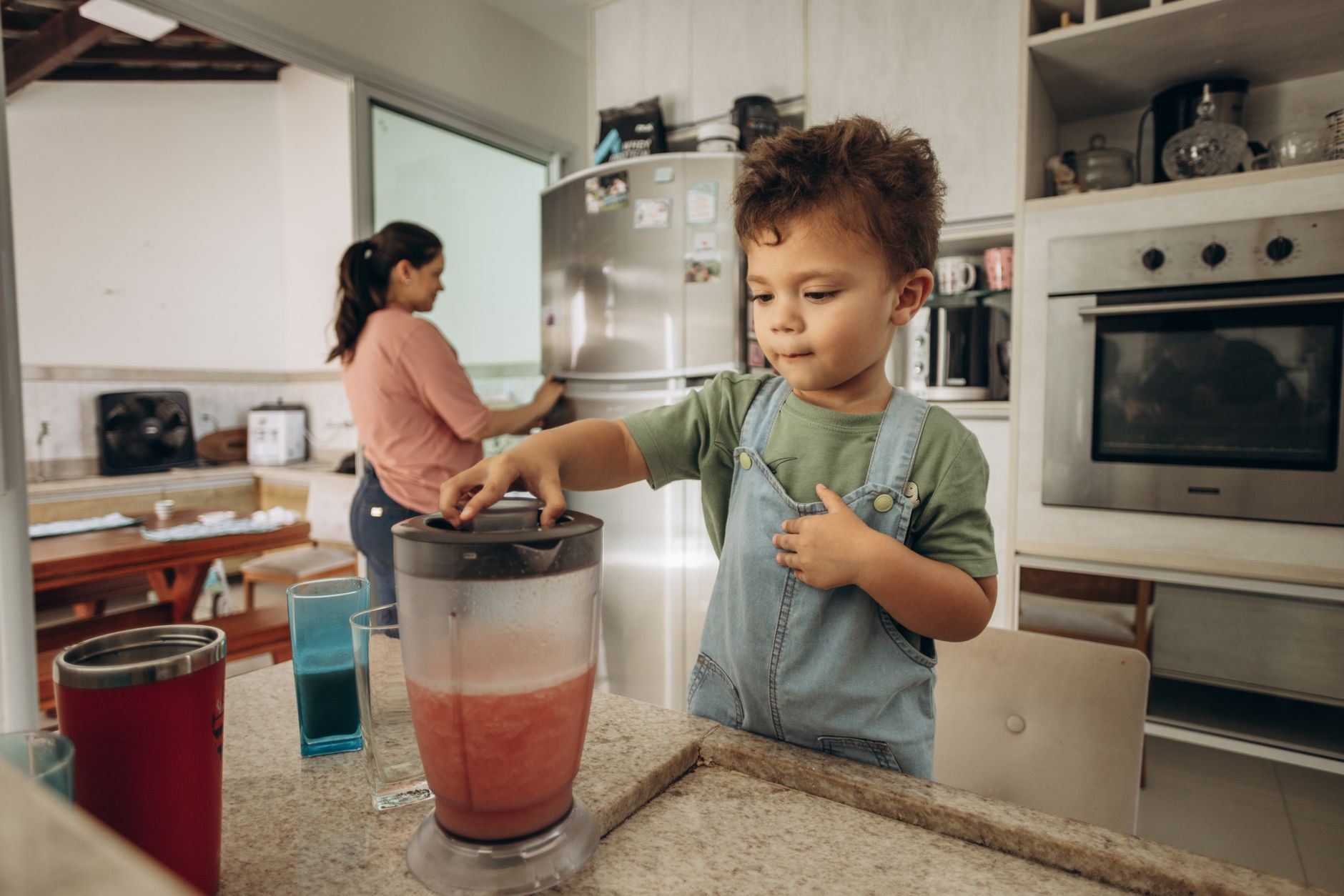 A young boy helps his mother make a smoothie using a blender in a modern kitchen.