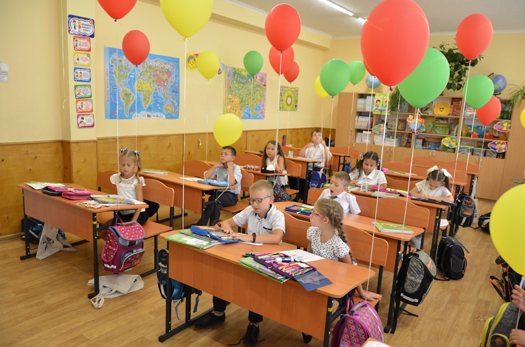 Children Sitting At Desks In A Classroom Decorated With Balloons RajrNwpgQ9U
